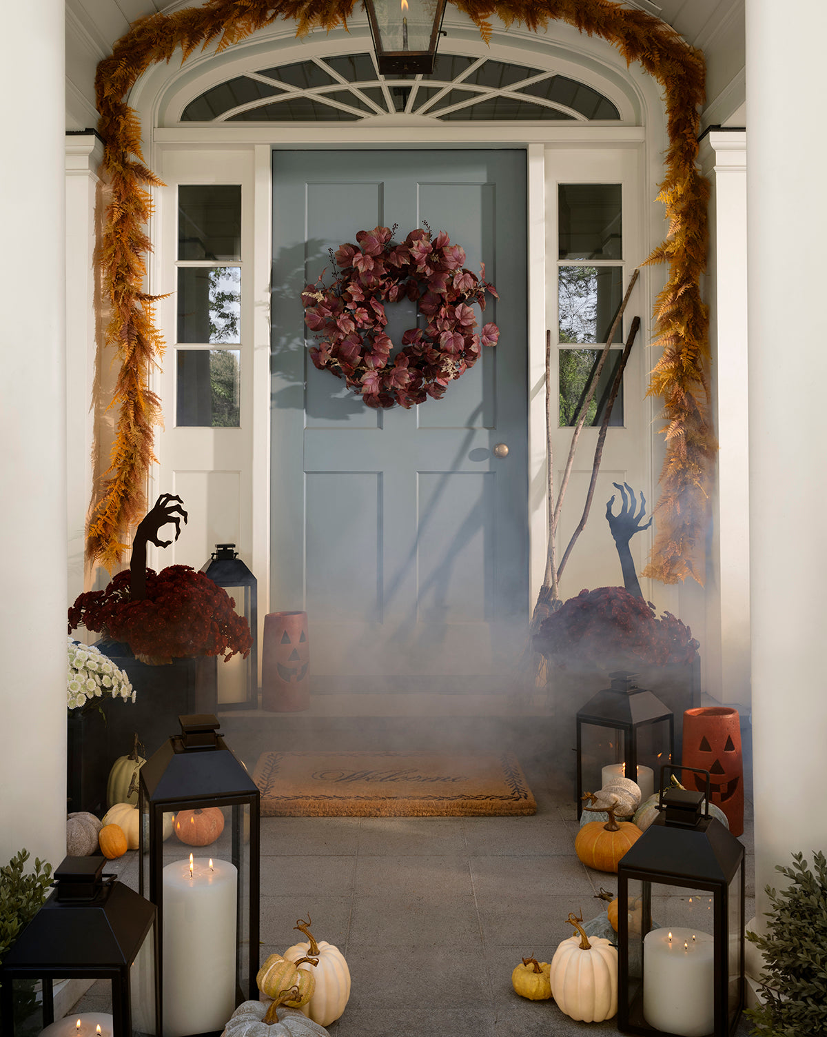 A front porch decorated for Halloween with pumpkins, lanterns, candles, a 6 Faux Golden Fern Garland, fake hands, a red wreath on the door, fog effects, autumn plants, jack-o-lanterns and spooky props for festive autumn style by McGee & Co.