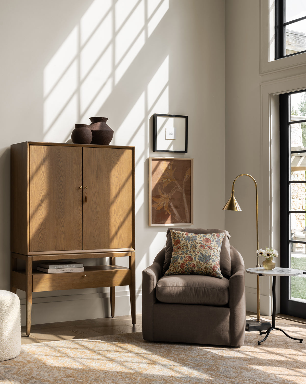 A cozy living room corner with a wooden cabinet, brown armchair and floral pillow, framed wall art, marble side table with flowers, sunlight streaming through tall windows, and the Vision Floor Lamp featuring a brass shade, by McGee & Co.
