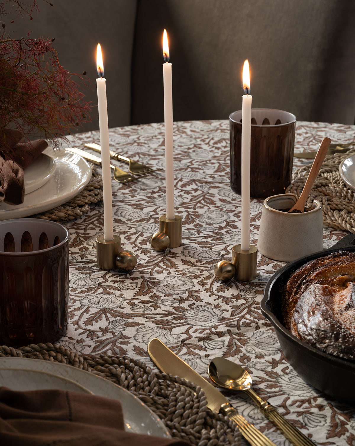A decorated dining table with a floral tablecloth, three lit white candles in gold holders, brown glass cups, gold cutlery, woven placemats, a cast-iron skillet of bread, and a Malco Salt Cellar with a wooden spoon.