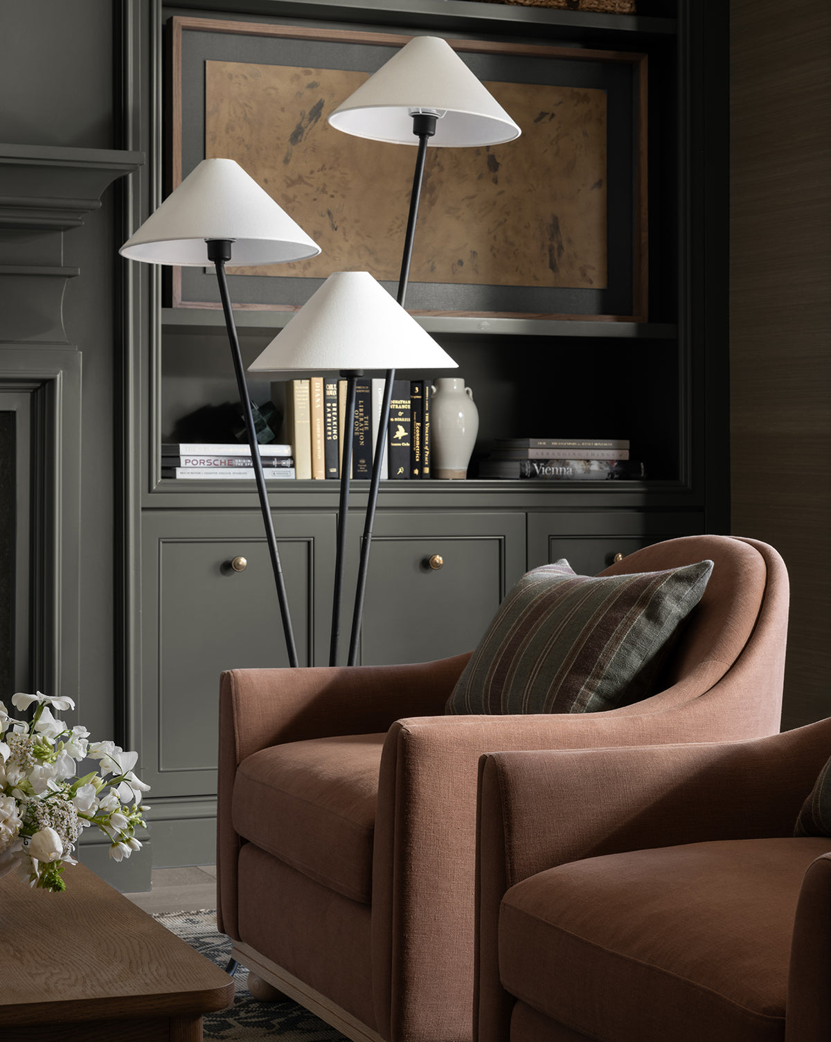 A cozy living room with a soft brown armchair, striped pillow, and the Cedar Floor Lamp with linen shades. Built-in dark green shelves behind hold books, a vase, and framed artwork. White flowers rest on a wooden table, by McGee & Co.