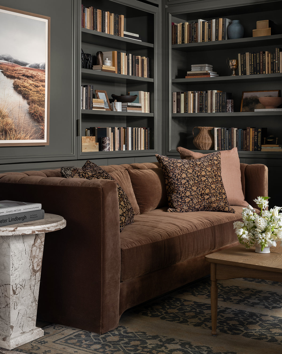 A cozy living room with a Makerspalm Agatha Sofa 90 in dark brown velvet, patterned pillows, built-in bookshelves filled with books, a marble side table, wooden coffee table with flowers, and a landscape photo on the wall by McGee & Co.
