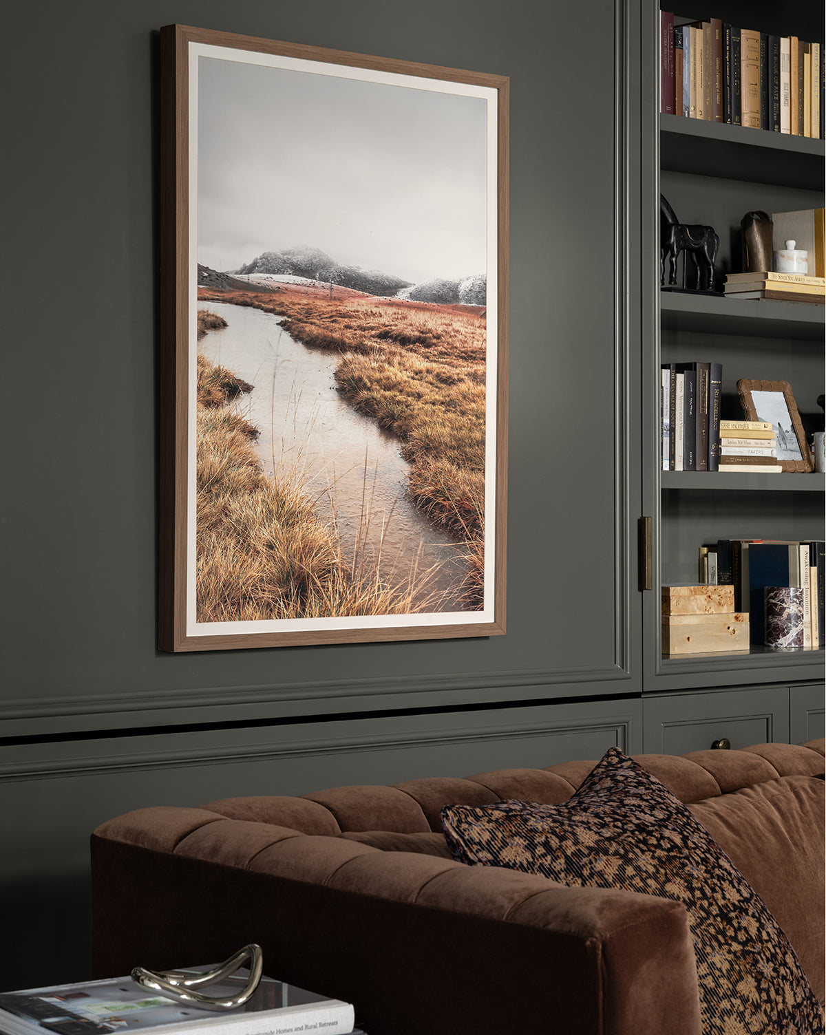 “Autumn Marshes” giclee print features a stream winding through autumn marshes and snowy mountains, shown in a walnut wood frame on a dark gray wall beside built-in shelving and a brown velvet sofa with patterned pillows by McGee & Co.