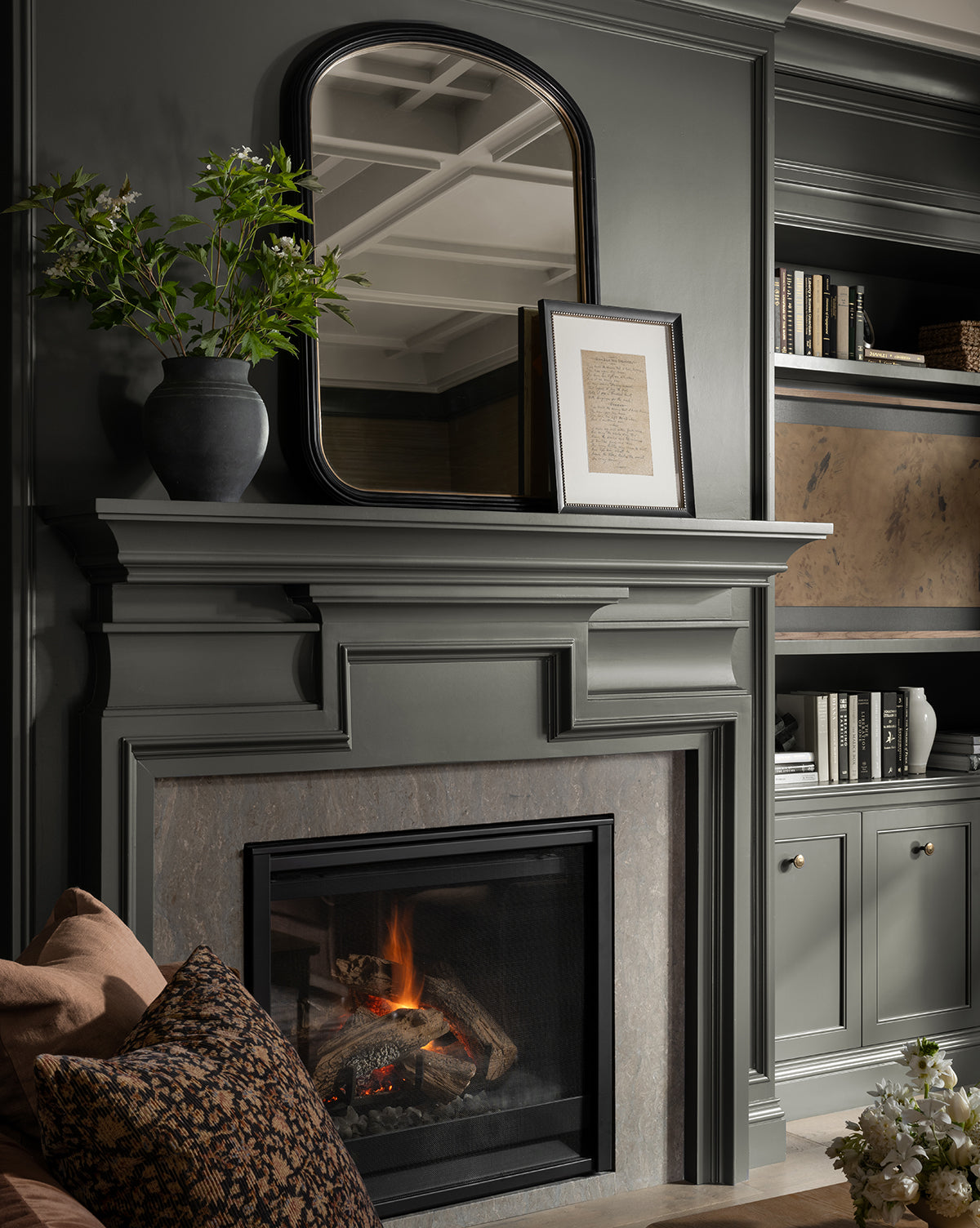 A cozy living room features a gray fireplace, glowing fire, black vase of greenery, an arched mirror, and framed handwritten song lyrics—Give Back the Dreams. Built-in shelves filled with books and decor complete the space.