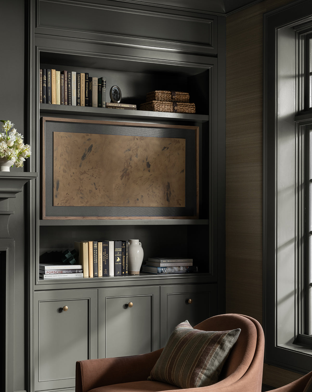 Dappled features a built-in bookshelf with dark gray cabinets, books, deckled edges art, and a TV with an artistic tan cover. Nearby sits a brown armchair with a striped pillow by a sunlit window, by McGee & Co.