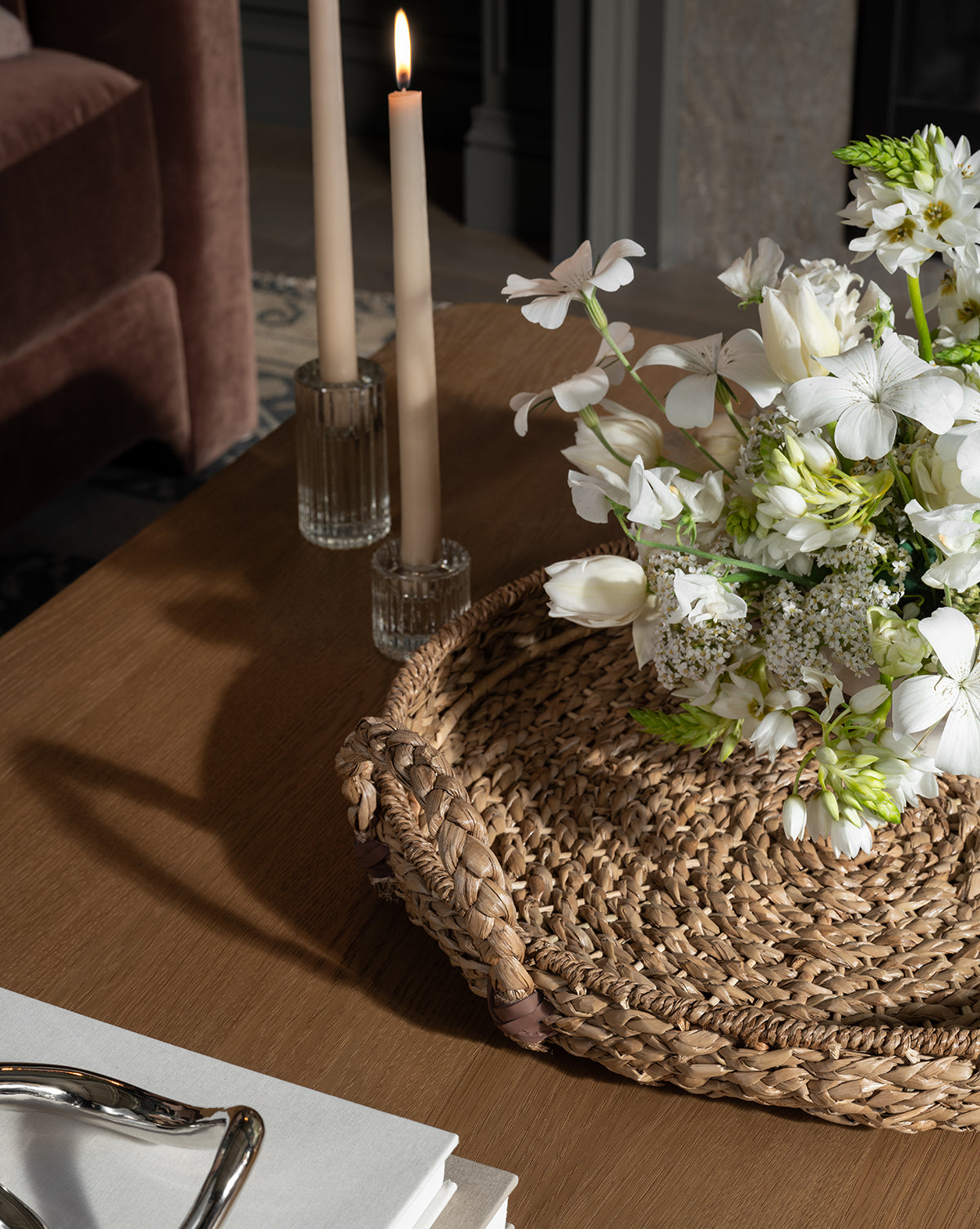 A Salter Woven Tray with a bouquet of white flowers rests on a wooden table beside two lit beige candles in glass holders. A brown chair and a stack of white books are also visible, by McGee & Co.