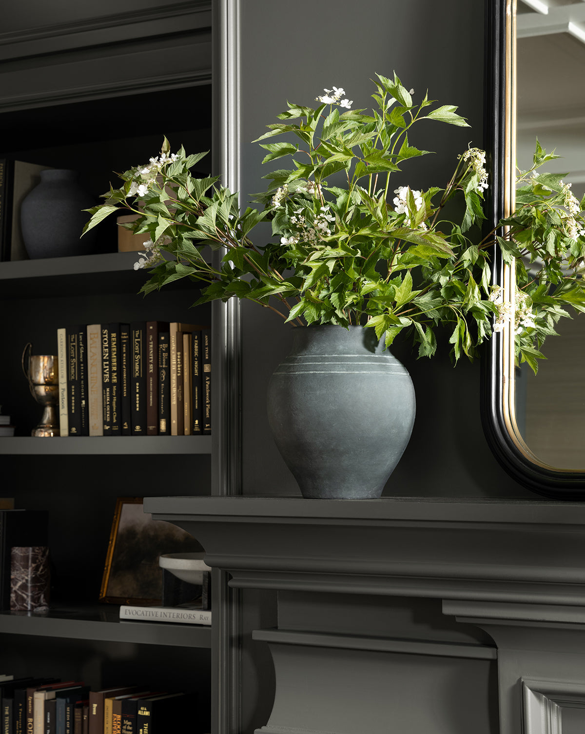 A dark gray room with built-in shelves, books, and the Ethel Vase—black terracotta filled with leafy branches and white flowers—on a mantel beneath a mirror, ideal for rustic home decor lovers, by McGee & Co.