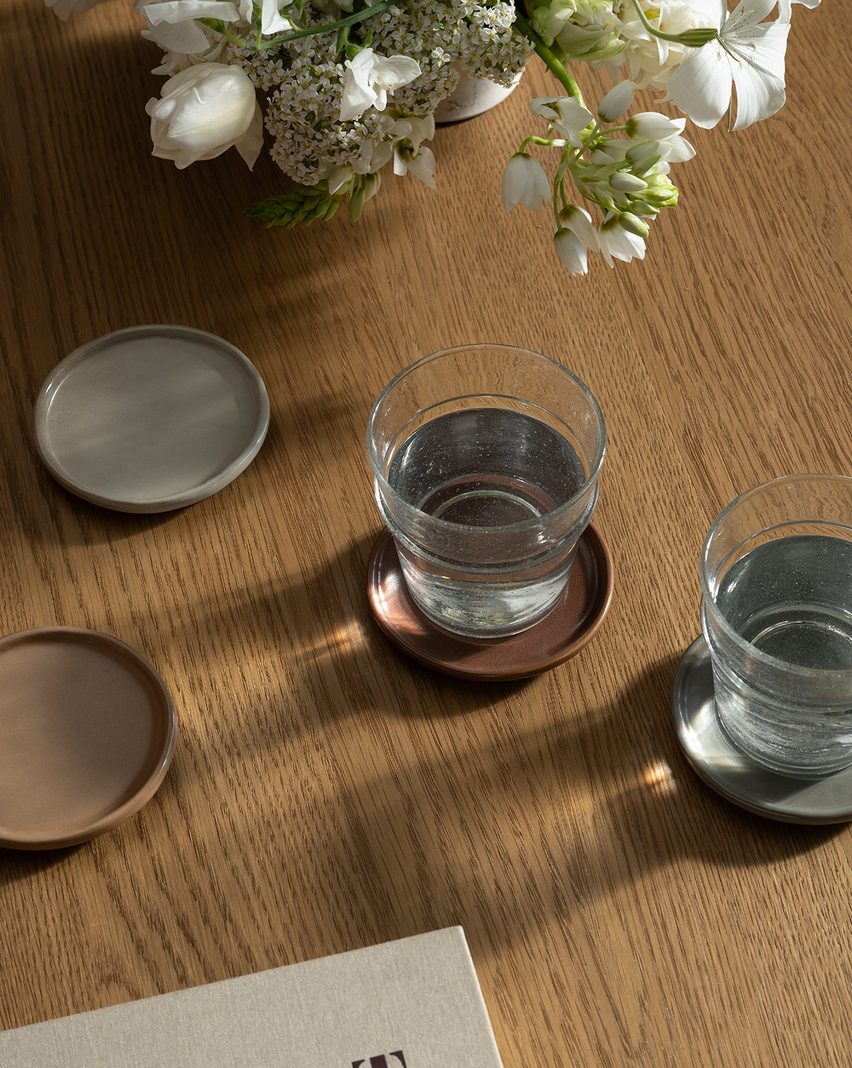 Two glasses of water rest on Speckled Ceramic Coasters (Set of 4) atop a wooden table, surrounded by empty coasters, a beige book, and white flowers in a vase. Sunlight creates soft shadows across this peaceful tabletop arrangement, by McGee & Co.