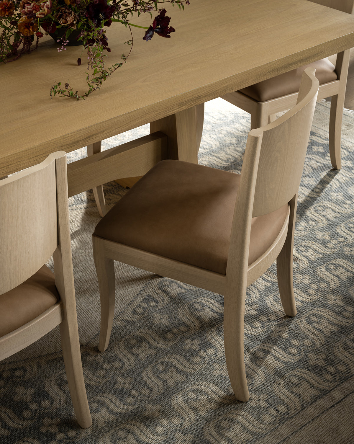 A light wooden dining table with Shepherd Dining Chairs, each featuring white oak frames and brown leather cushions, sits on a patterned rug. A floral arrangement is partially visible at the top of the image, by McGee & Co.
