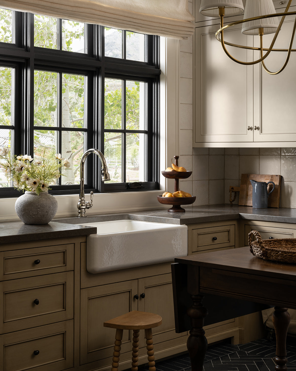 A cozy kitchen features a farmhouse sink beneath black-trimmed windows, beige cabinets, and a wooden stool. A vase with the Cream Cosmos Stem sits on a dark wood table, alongside a wicker tray and tiered fruit stand as sunlight streams in, by McGee & Co.