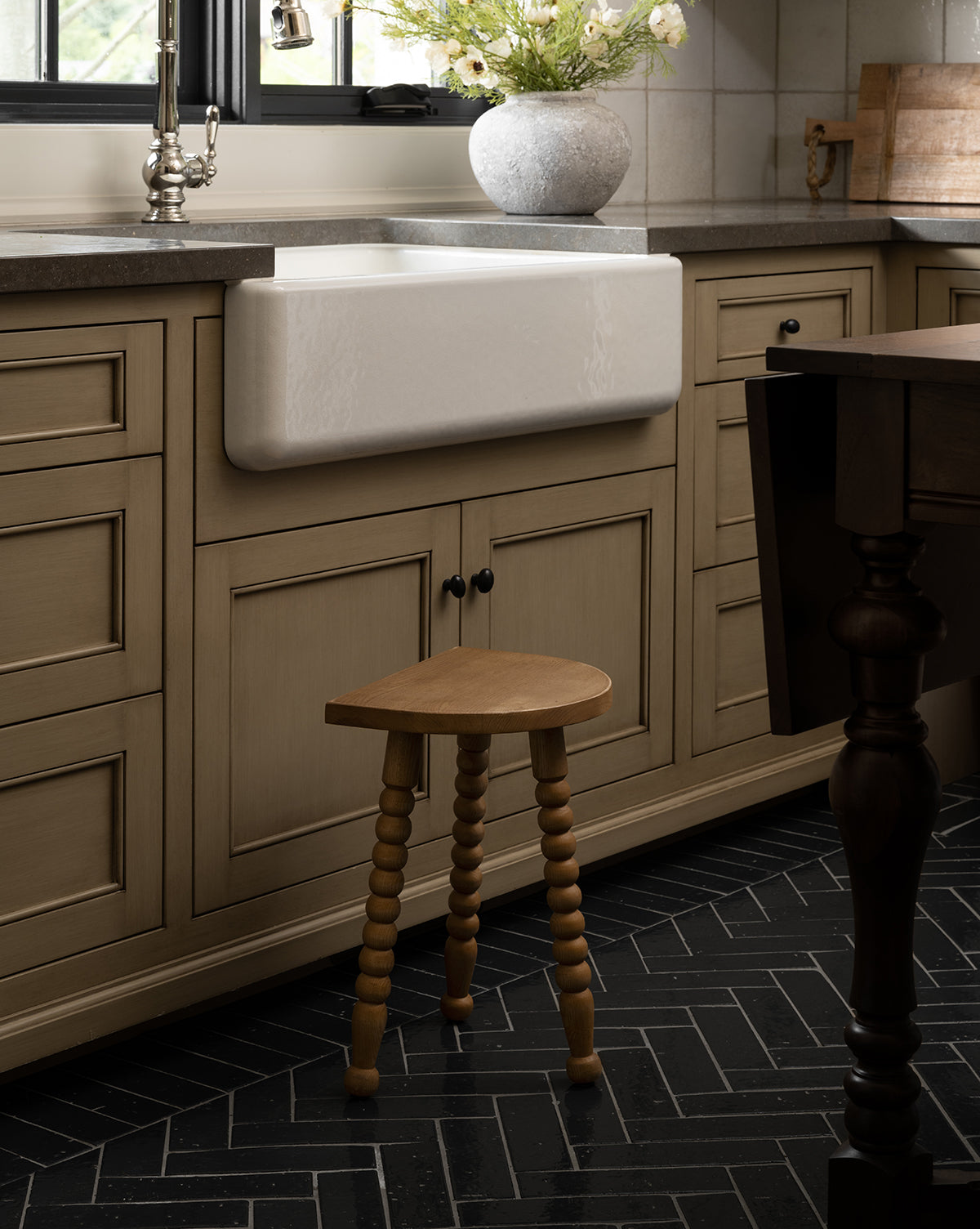 The Makerspalm Alice Honey Wood Stool with spindle legs sits before a farmhouse sink and beige cabinets in a kitchen featuring black herringbone tile flooring. A vase of flowers decorates the countertop by the window, McGee & Co.