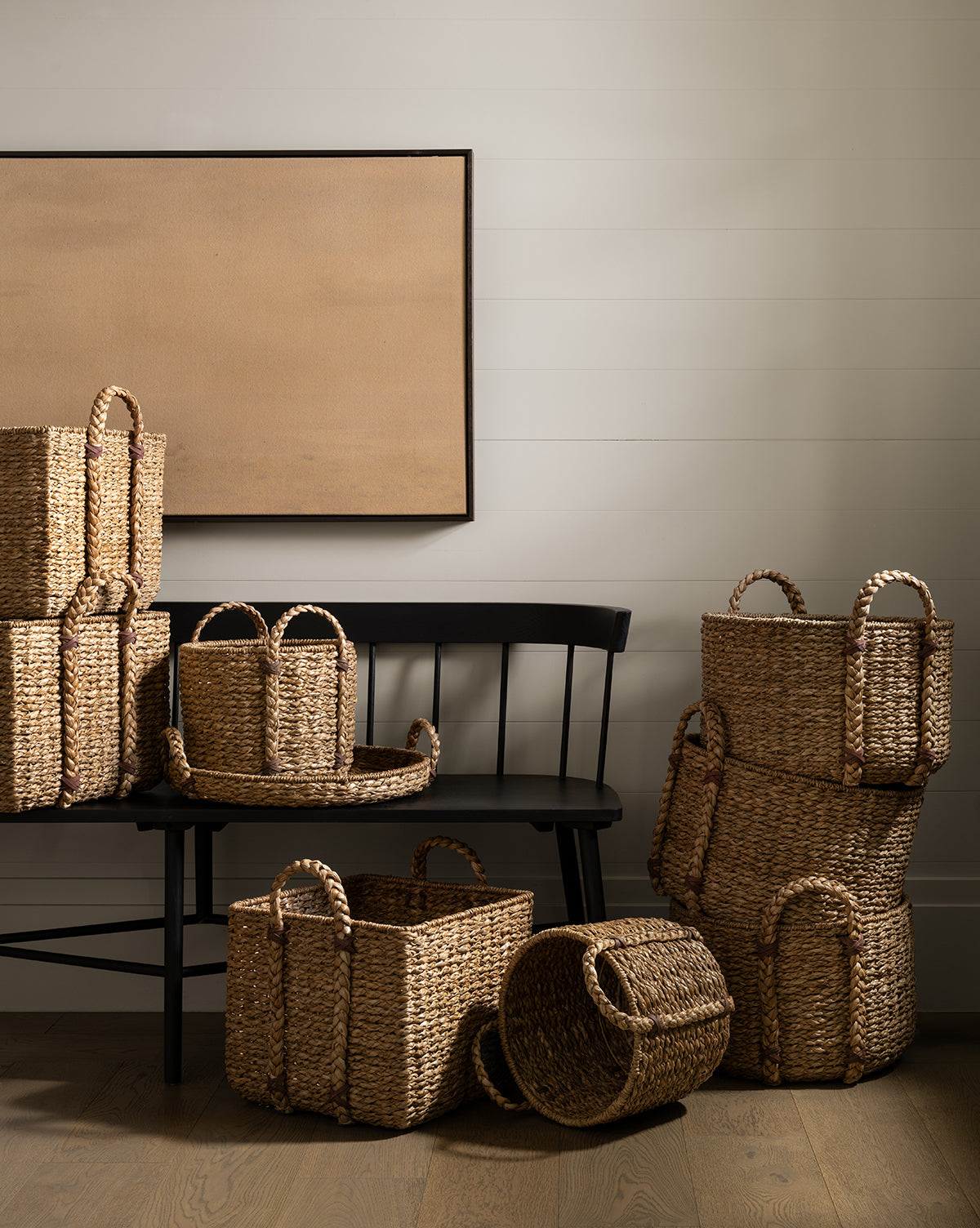 The Salter Woven Square Basket, along with other handled woven baskets, is displayed on and around a black wooden bench set against a white shiplap wall and beige canvas, adding an organized rustic touch to the wooden-floored space, by McGee & Co.