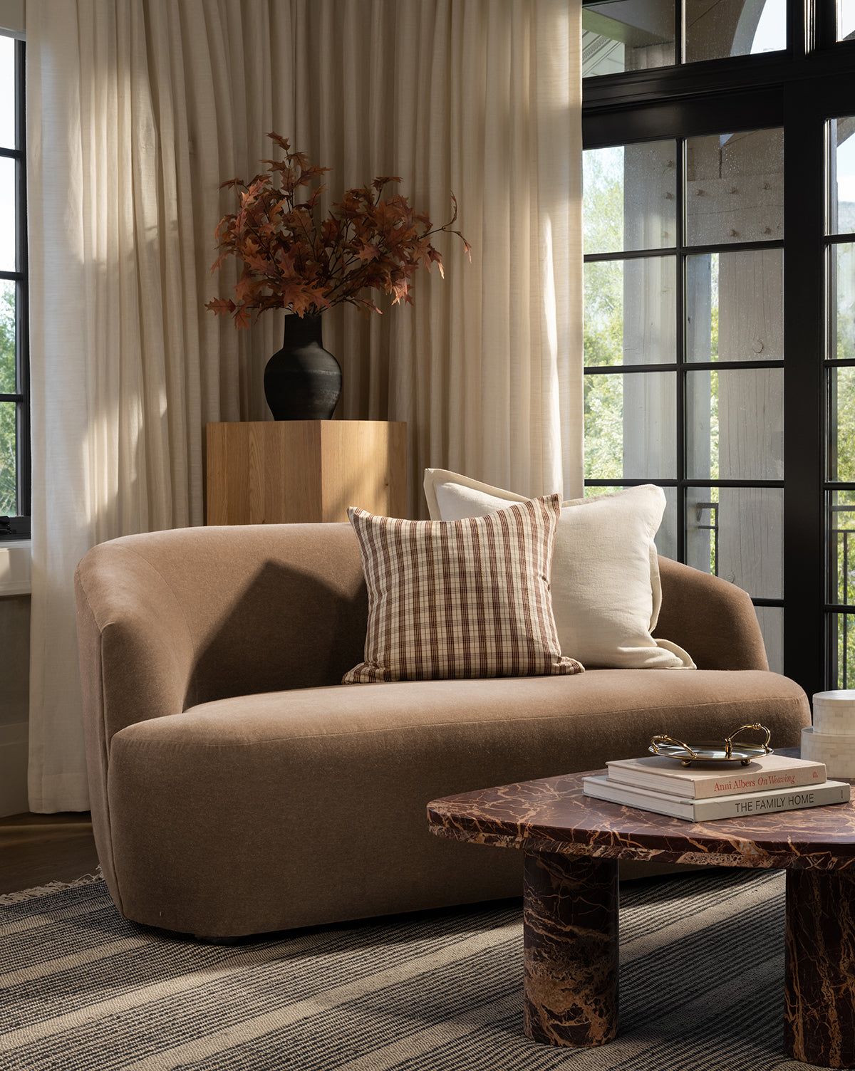 A modern living room with a curved brown sofa, plaid and white cushions, a wooden side table topped with a black vase of autumn leaves, a marble coffee table, large windows with cream curtains, and a decorative Liam Double Flange Pillow Cover for added charm, by McGee & Co.