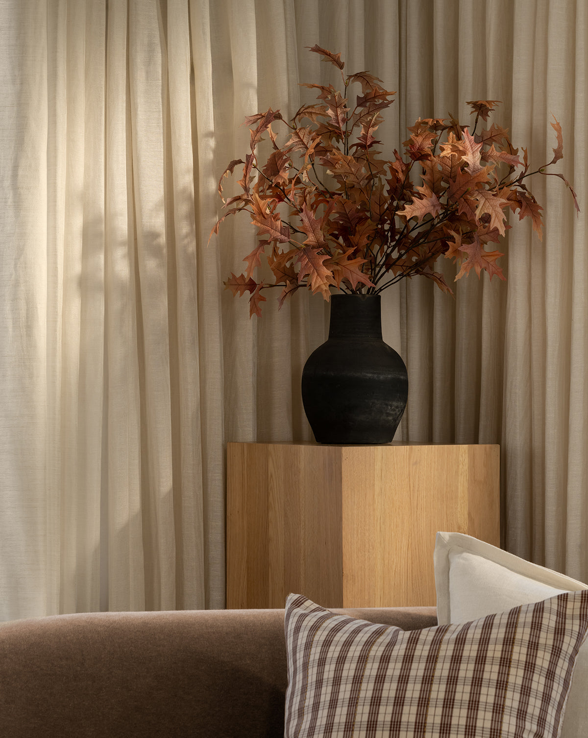 A Faux Autumn Oak Stem adds a touch of autumn to a black vase on a wooden pedestal, set against beige curtains. In front, a brown sofa with plaid and cream pillows completes the warm, cozy atmosphere.