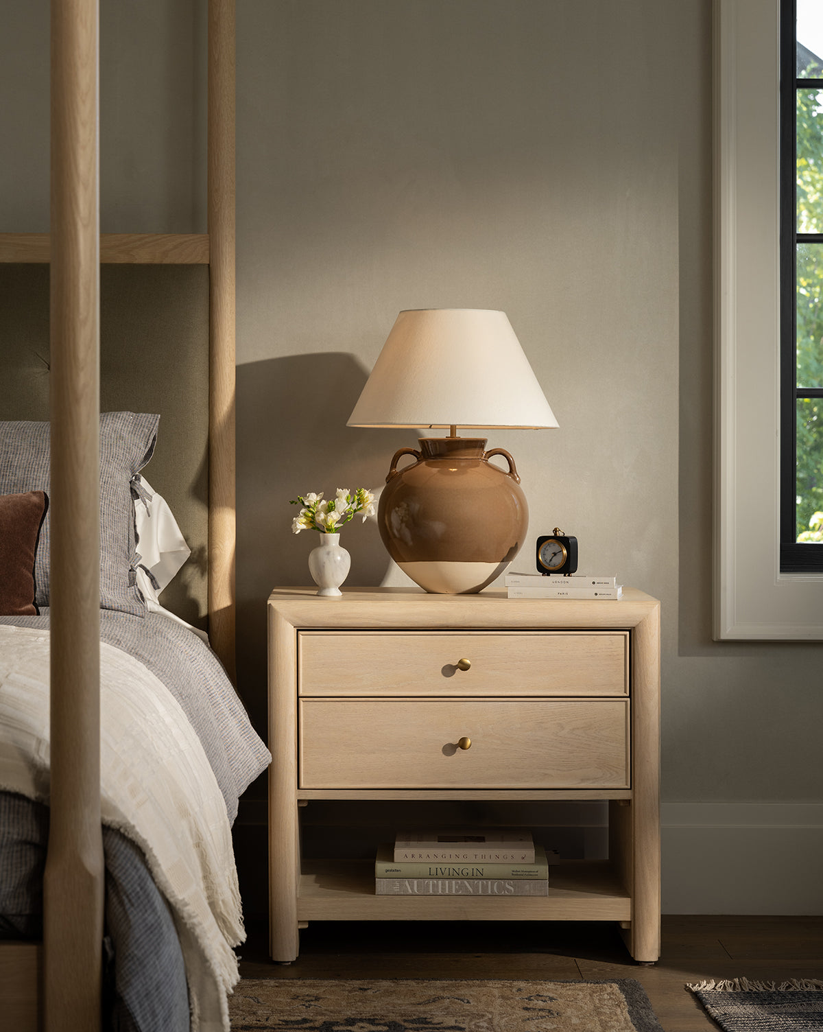 A Tellerbeck Nightstand sits by the bed, featuring two drawers and topped with a brown lamp, white vase of flowers, and black alarm clock—ideal for stylish bedroom organization as sunlight filters in by McGee & Co.