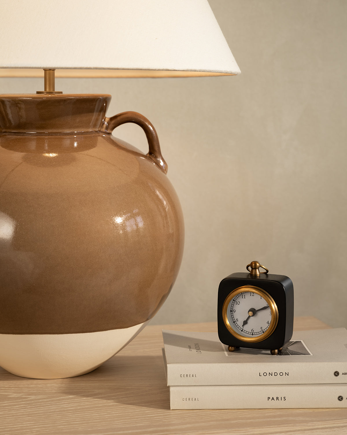 A brown ceramic lamp and the Kya Clock sit on books labeled Cereal, London, and Paris atop a light wooden nightstand against a beige wall, by McGee & Co.