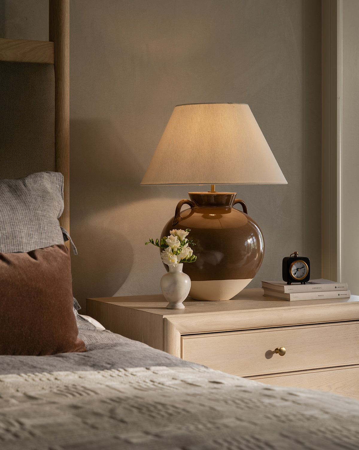 A cozy bedside scene features the Amphora Table Lamp, a vase of white flowers, books, and a black clock on a light wood nightstand beside a bed with neutral bedding by McGee & Co.