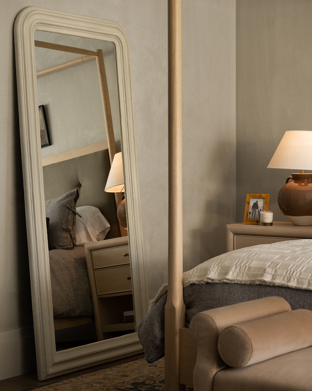 A cozy bedroom features a Breslin Floor Mirror reflecting the bed, nightstand, and lamp. A beige knit blanket, tan leather bench, and brown lamp with a photo frame sit on the nightstand near plaster-finished walls by McGee & Co.
