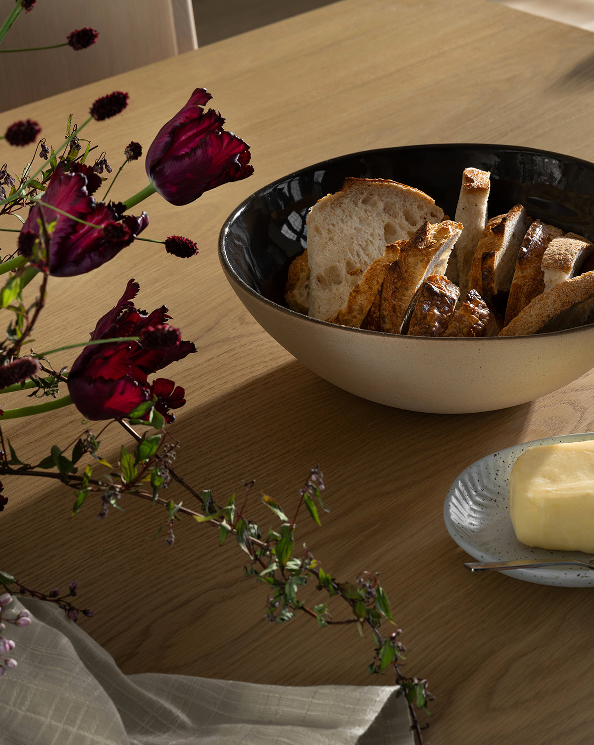 The Joanne Serving Bowl, filled with sliced artisan bread, sits on a wooden table next to butter, dark red flowers, and a folded napkin by McGee & Co.