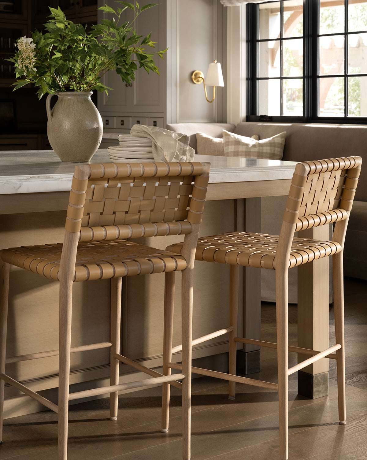 Two Greely Stools with a white oak frame sit at a light marble kitchen island. A beige ceramic pitcher with green leafy branches decorates the countertop as sunlight streams through black-framed windows, by McGee & Co.
