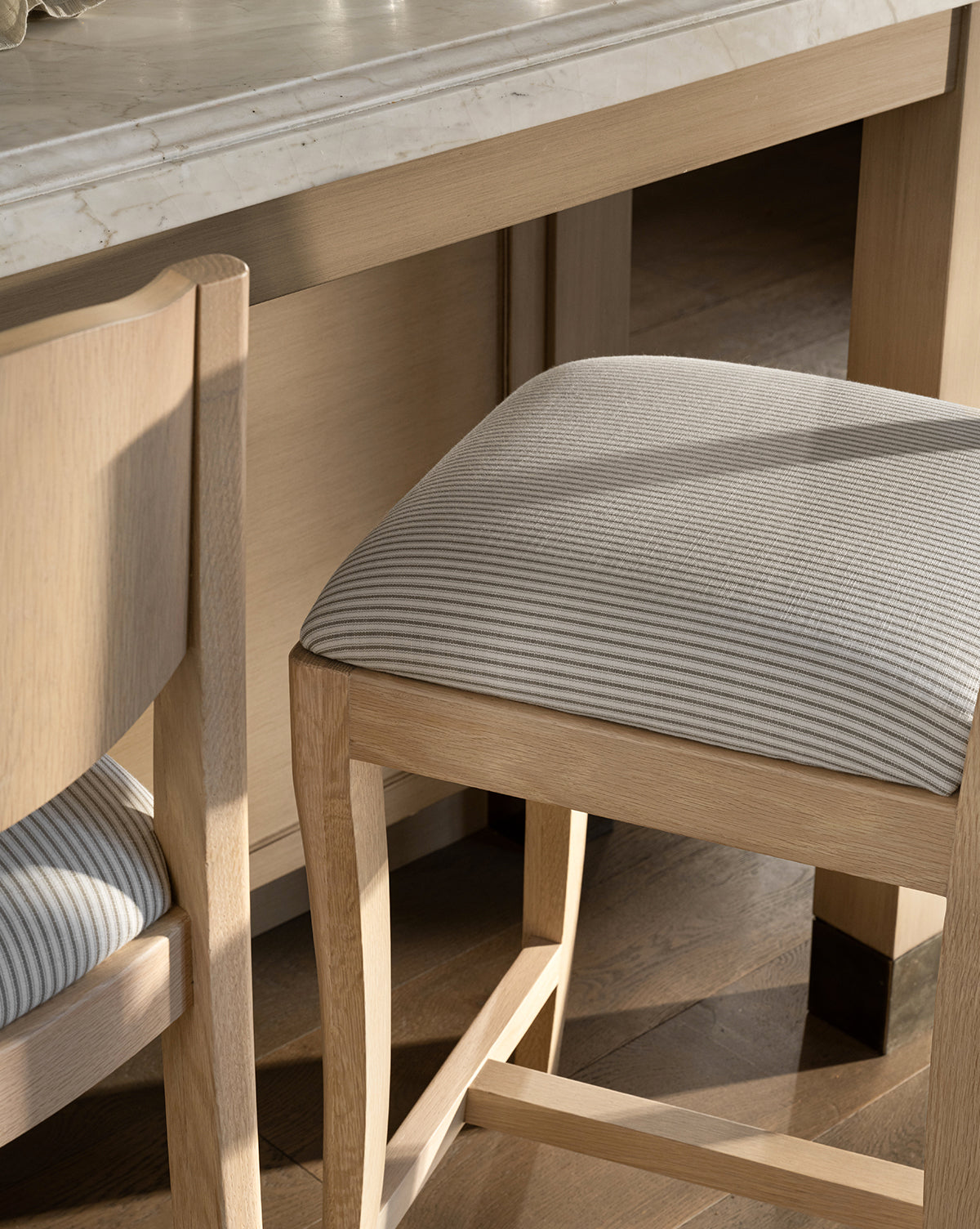A close-up of the Makerspalm Shepherd Counter Stool featuring gray and white striped performance upholstery, set by a marble-top table on a wooden floor in a sunlit room. McGee & Co.