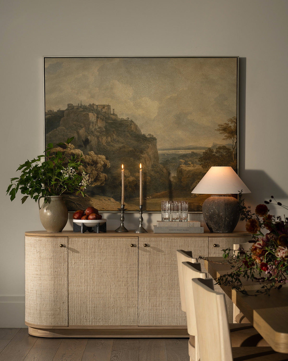 The Linu Woven Sideboard is styled with a large landscape painting, two lit candles, a lamp, a vase of greenery, fruit bowl, and glasses. To the right, a dining table with chairs and a floral arrangement is visible by McGee & Co.