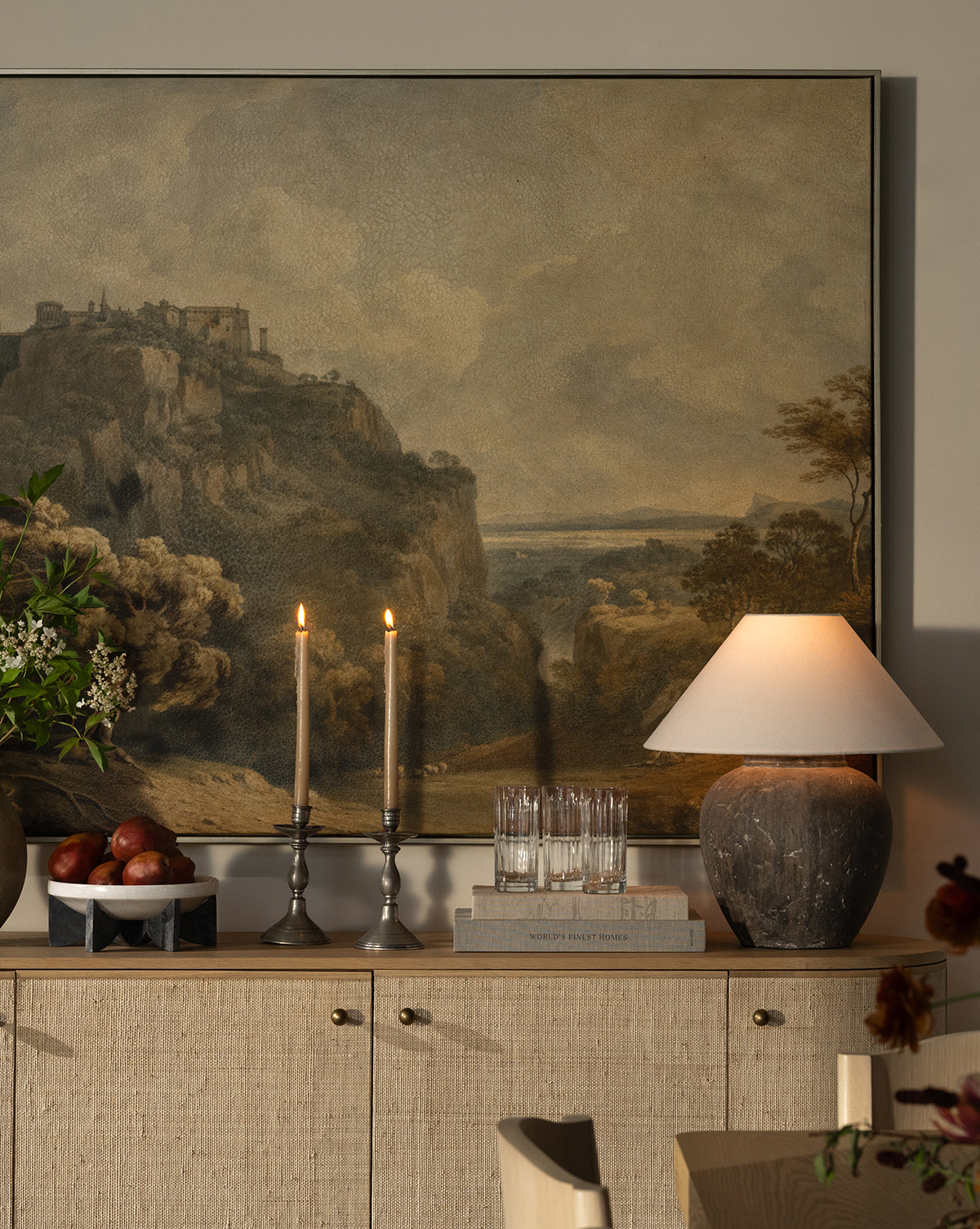 A cozy sideboard holds a rustic table lamp, two lit candles, stacked books, apples in a bowl, glasses, and a vase of flowers beneath the dramatic Overlooking Tivoli landscape painting of a cliffside village by McGee & Co.