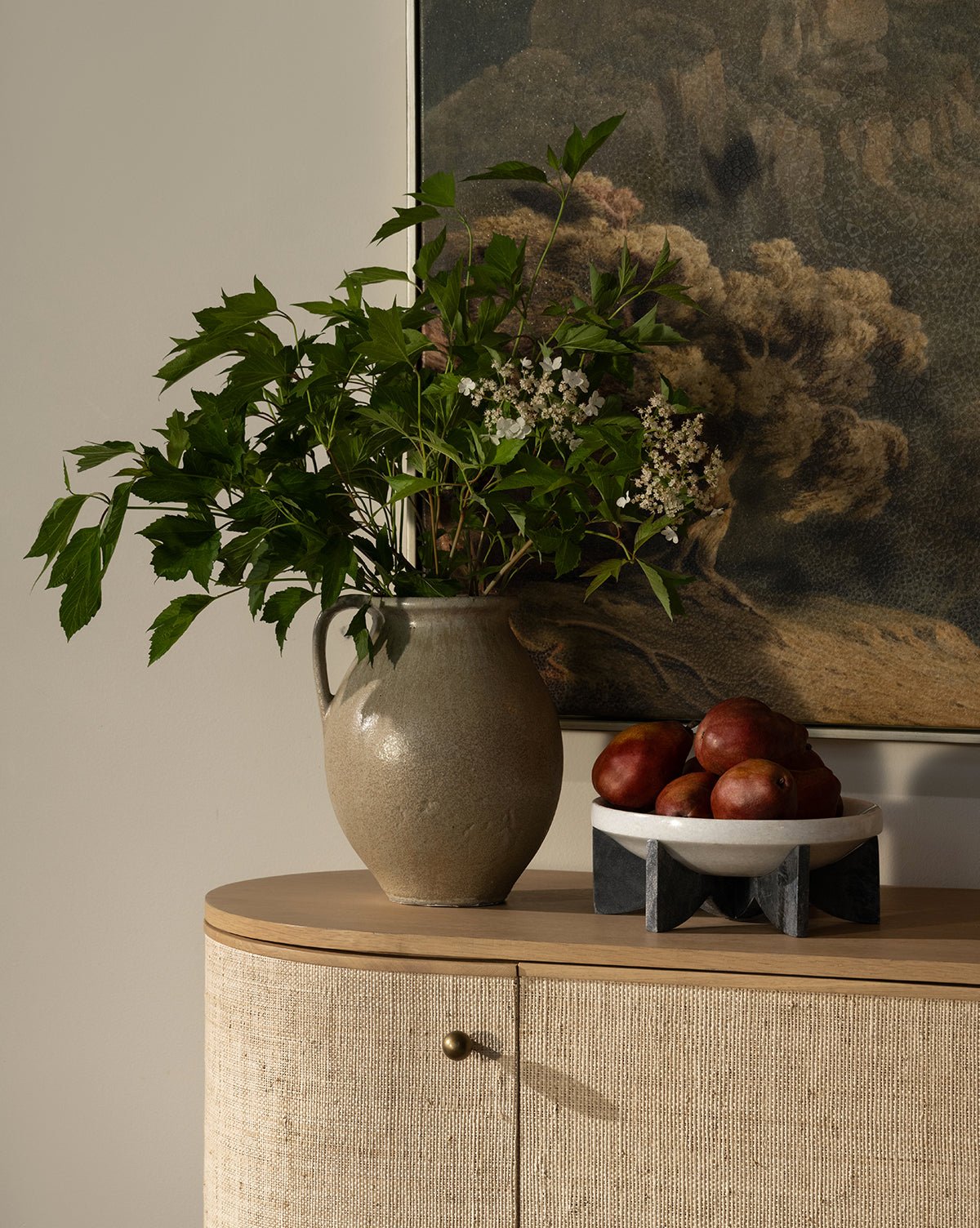 A stoneware jug with green leafy branches and white flowers sits on a light wood cabinet next to the Mahoney Bowl filled with red apples, by McGee & Co. A landscape painting hangs on the wall in the background.
