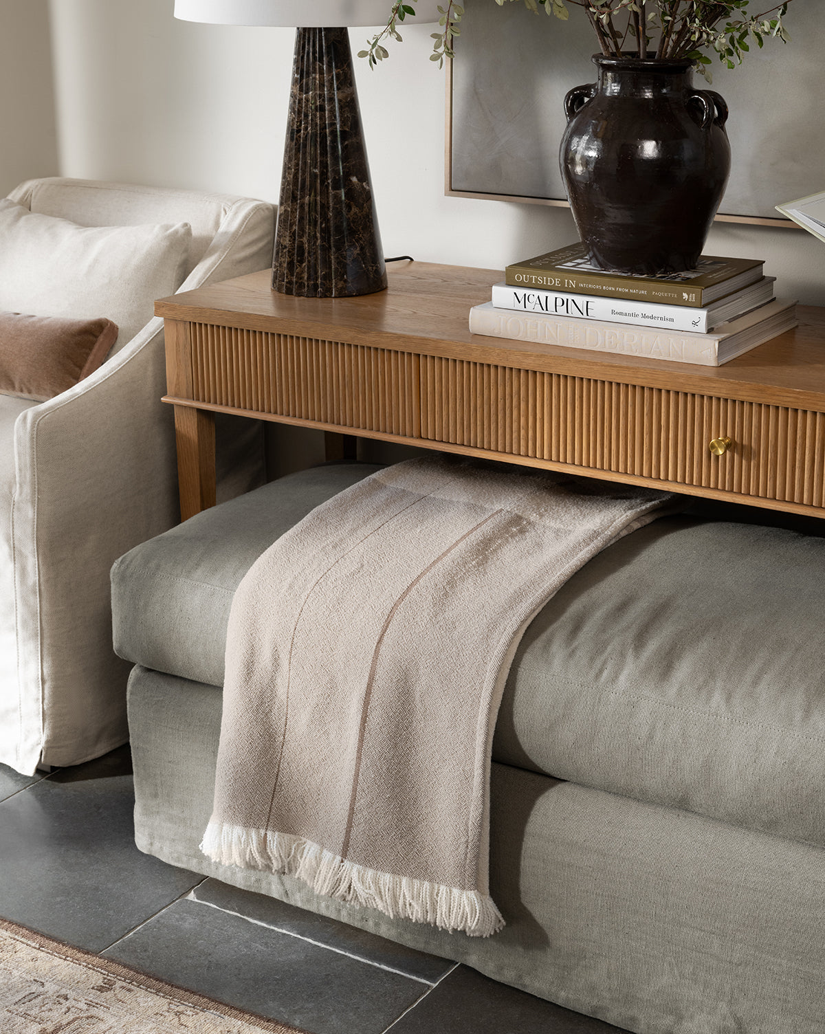 A cozy neutral-toned living room corner features a wooden console table with a dark vase, books, and a marble lamp. Beneath it, a cushioned bench displays the Alex Plaid Throw by ILA HOME FASHIONS in light beige for added texture. Styled by McGee & Co.