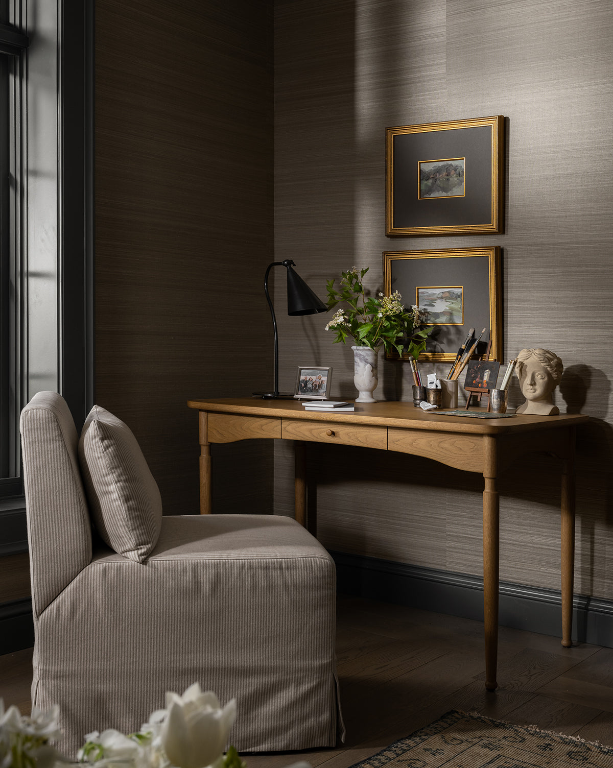 A cozy corner workspace features a wooden desk, beige upholstered chair, Lupe Table Lamp, potted plant, framed wall art, and desk accessories, all set against neutral-toned walls with a nearby window by McGee & Co.