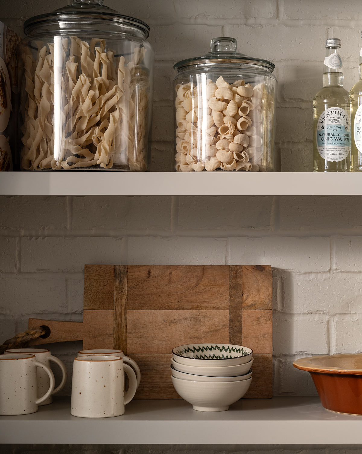Two glass jars of pasta and bottles sit on an upper shelf; below, ceramic mugs, a wooden cutting board, stacked bowls including the Mattia Hand-Painted Bowl with floral patterns, and a ceramic dish rest against a white brick wall by McGee & Co.