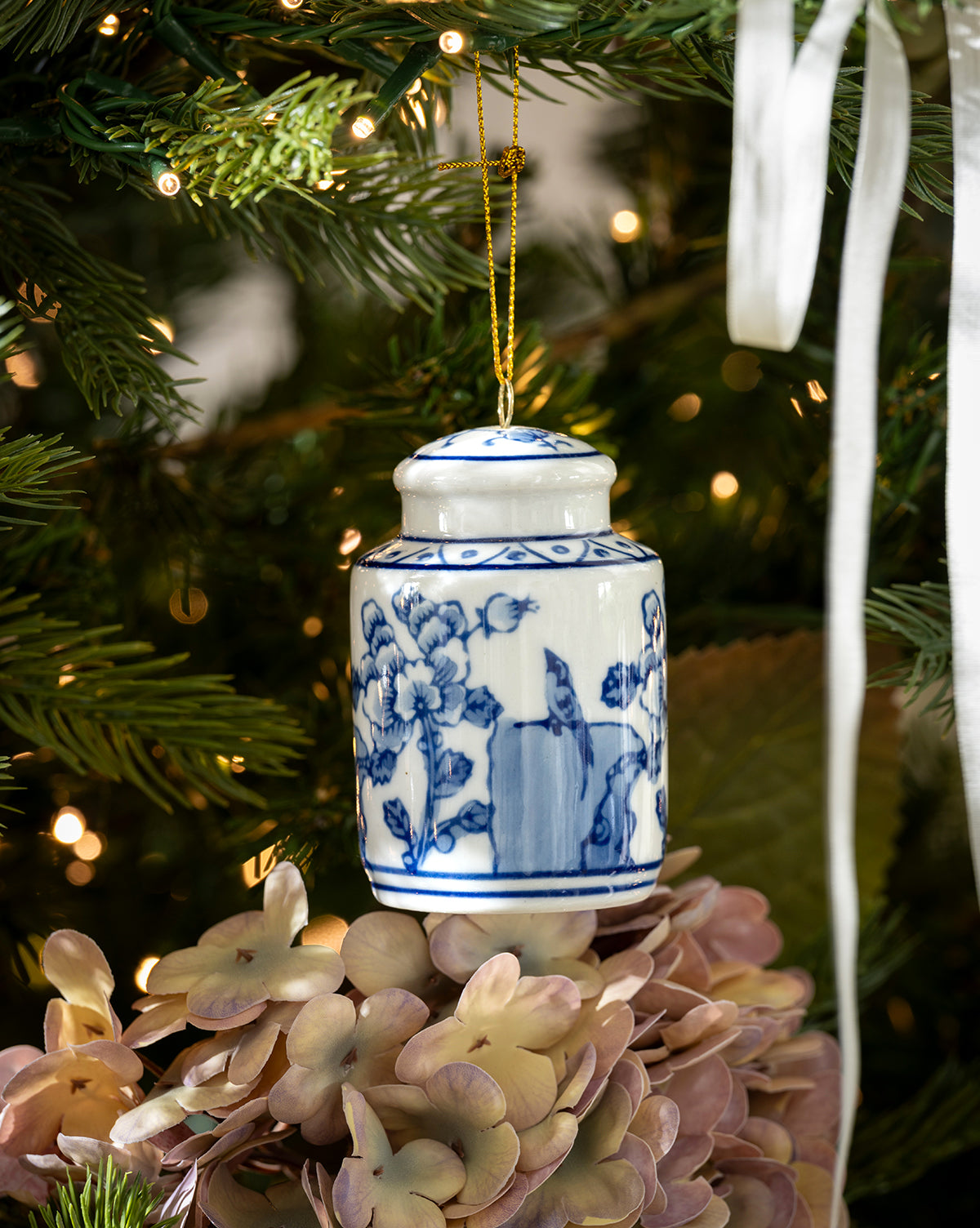 A blue and white floral porcelain jar ornament hangs on the Chateau Noel Tree, surrounded by greenery, warm lights, pale pink hydrangeas, and a luxurious velvet tree skirt, by McGee & Co.
