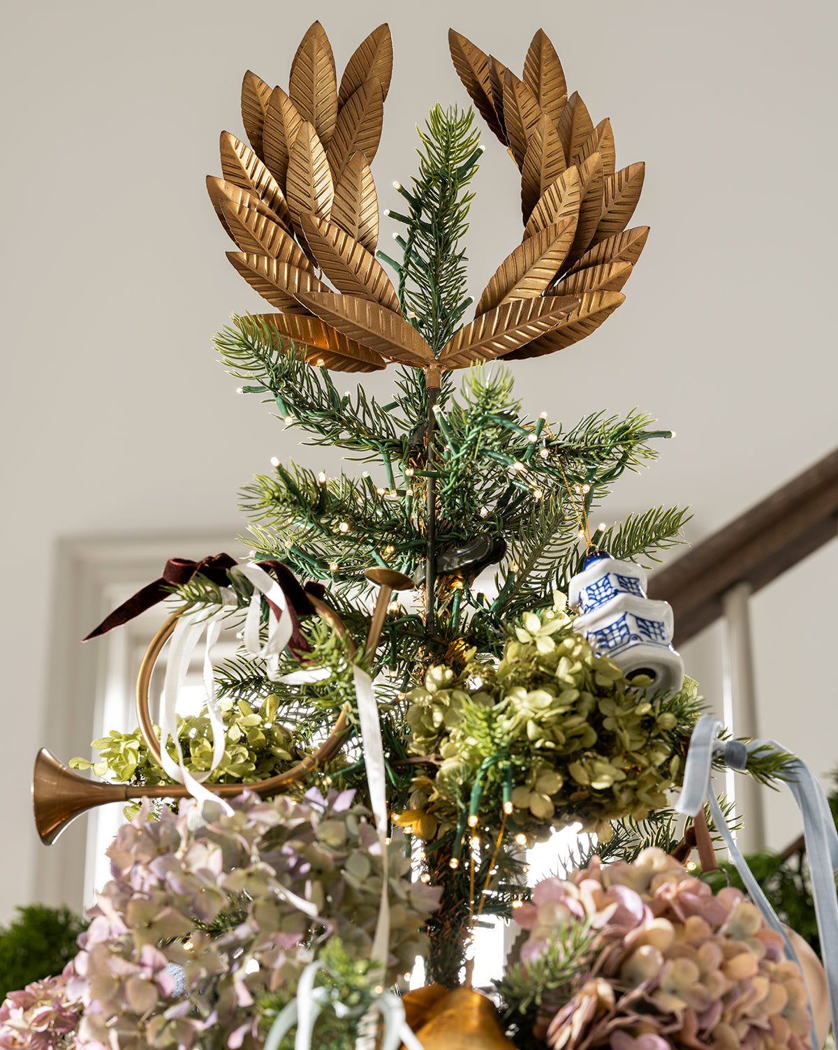 The Chateau Noel Tree is topped with gold leaves and adorned with dried flowers, white and blue ceramic ornaments, and a small gold horn. It stands in a chinoiserie planter with soft natural light in the background, by McGee & Co.