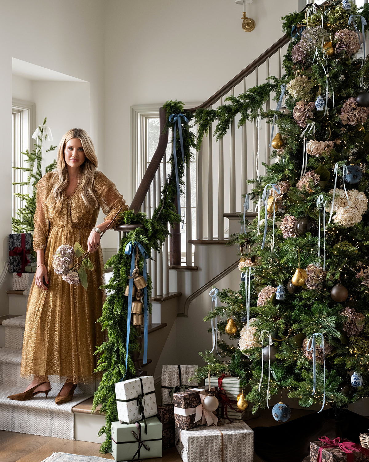 A woman in a gold dress stands by a staircase draped with garland, beside the Chateau Noel Tree decorated with ornaments and ribbons. Wrapped gifts sit on a velvet tree skirt in the bright, festive room, by McGee & Co.