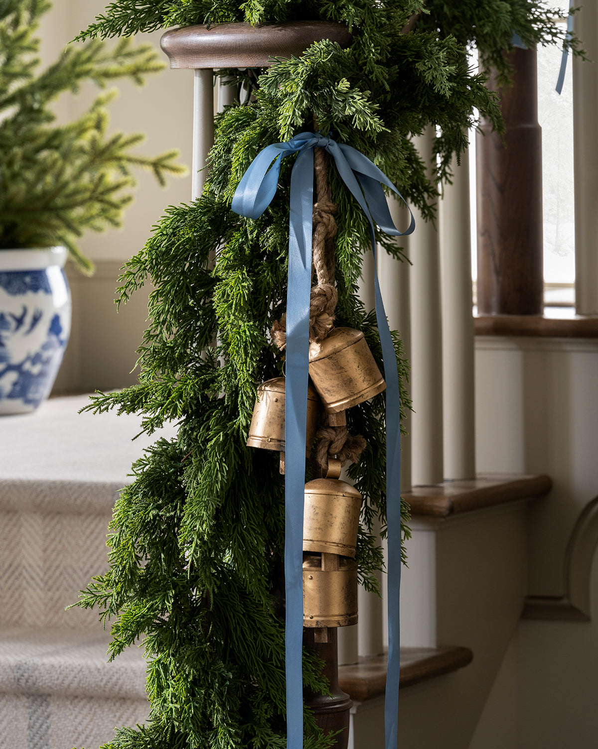 A staircase is adorned with lush green garland, gold Hanging Iron Bells, and a blue ribbon. In the background, a blue and white pot holds a small evergreen tree, creating festive holiday décor, by McGee & Co.