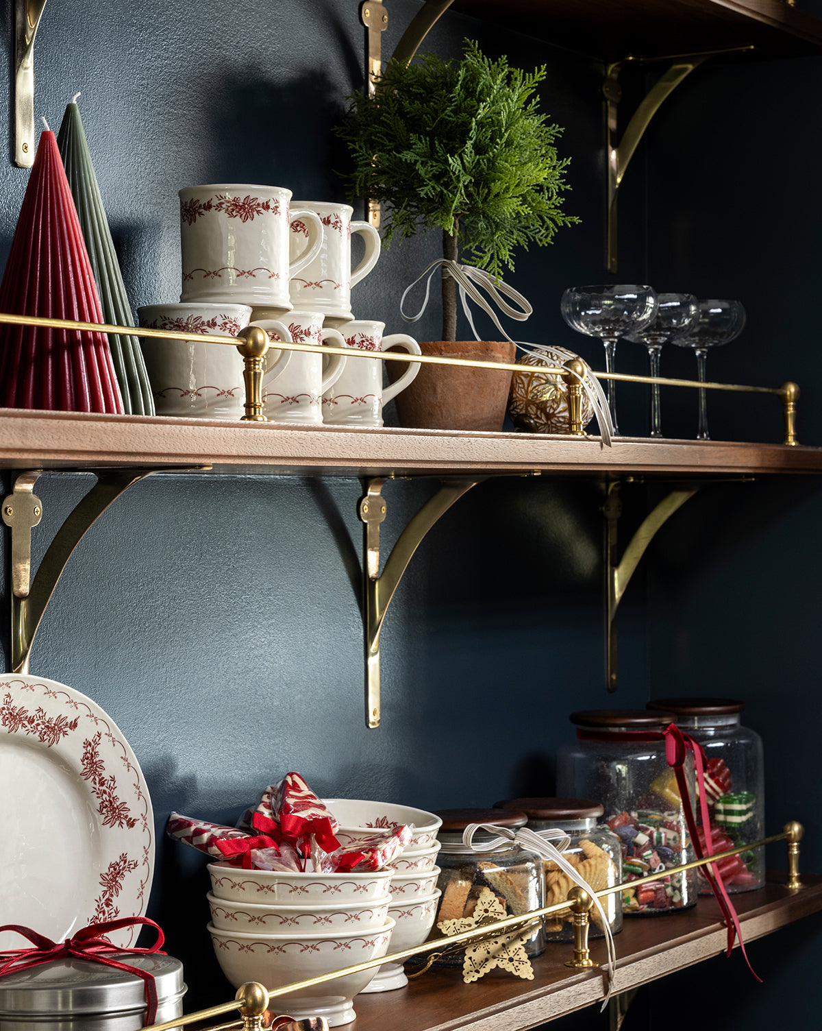 Wooden shelves with gold brackets hold stacked red-patterned mugs, bowls, plates, wine glasses, wrapped cookies, the 15.5" Cypress Faux Potted Topiary in a terracotta pot, red candles, and festive kitchen decor against a dark blue wall, by McGee & Co.