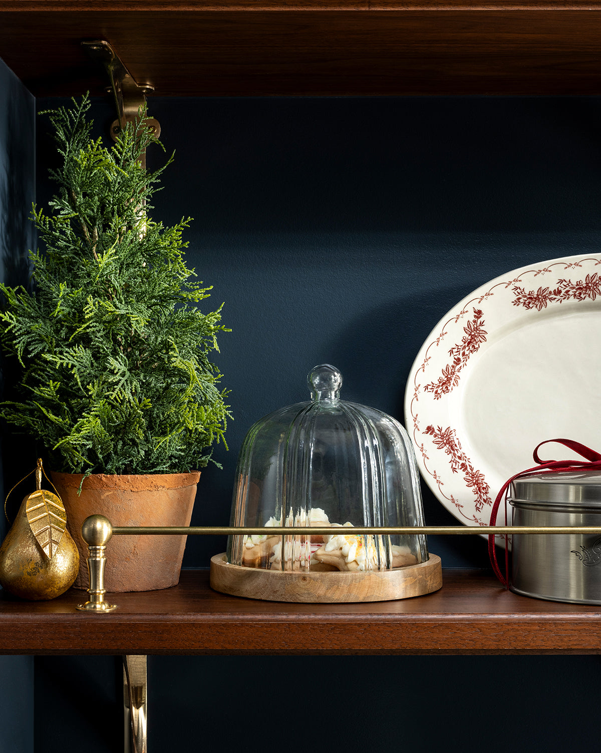 A 17" Cypress Faux Potted Tree, a golden pear ornament, a cheese plate with glass dome, a red-ribboned tin, and a white platter with red border are arranged on a wooden shelf against a dark wall, by McGee & Co.