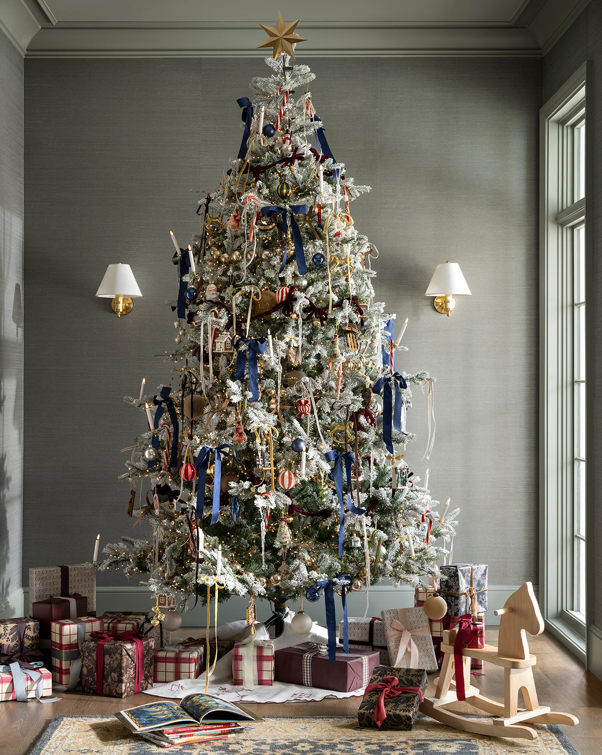 The Heritage Holiday Tree, topped with a gold star and decorated with blue ribbons and glass baubles, stands in a room filled with wrapped gifts, a wooden rocking horse, toys at its base, and large windows set against gray walls, by McGee & Co.