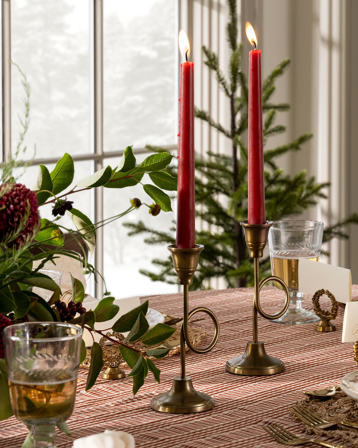 Two lit red taper candles in French Horn Taper Holders sit on a patterned tablecloth beside green foliage, a wine glass, and holiday decor, with festive styling and a window with a small Christmas tree in the background, by McGee & Co.