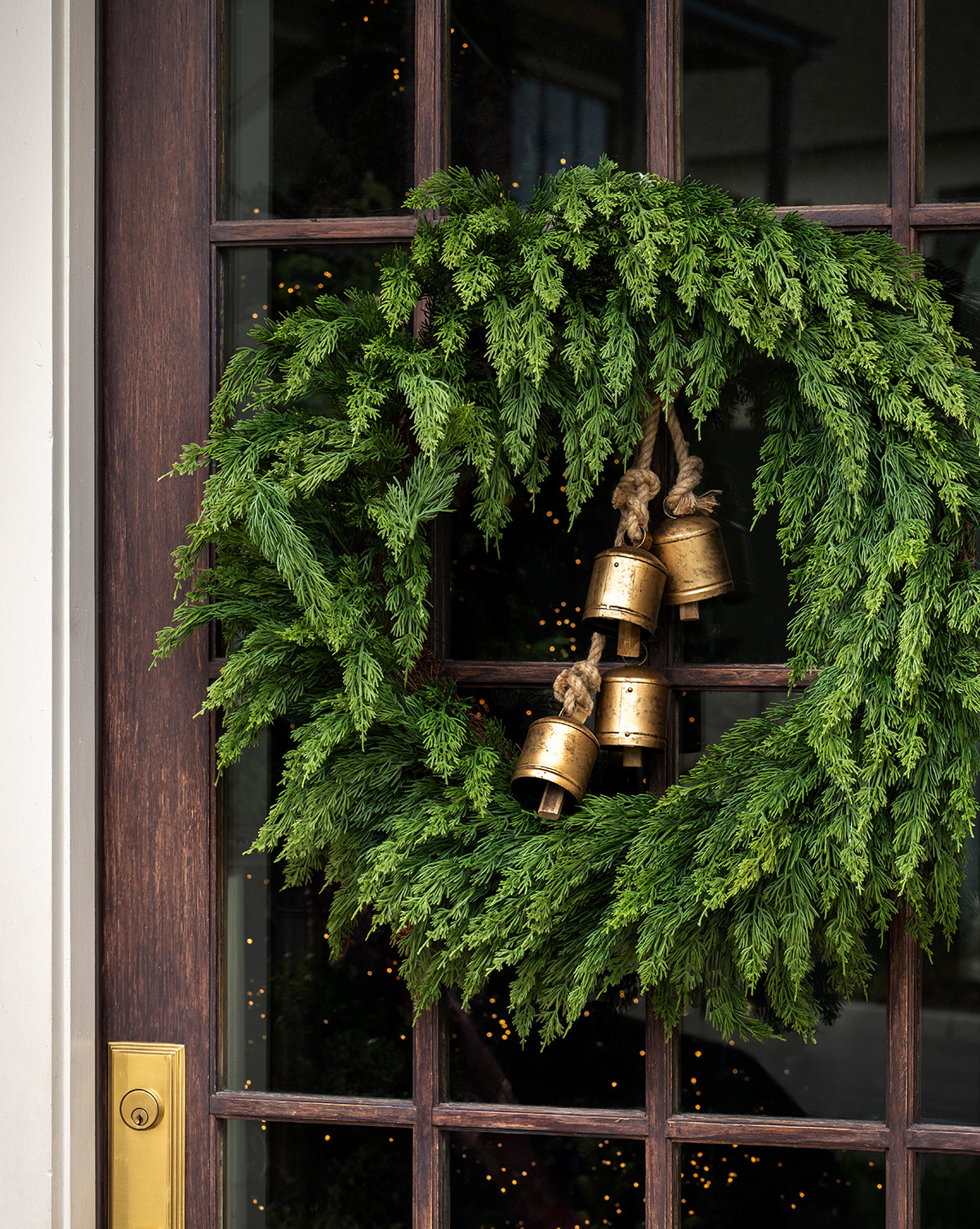 A festive green wreath featuring Hanging Iron Bells with a gold finish decorates a wooden door with glass panels, reflecting the warm glow of a Christmas tree and creating elegant holiday décor, by McGee & Co.