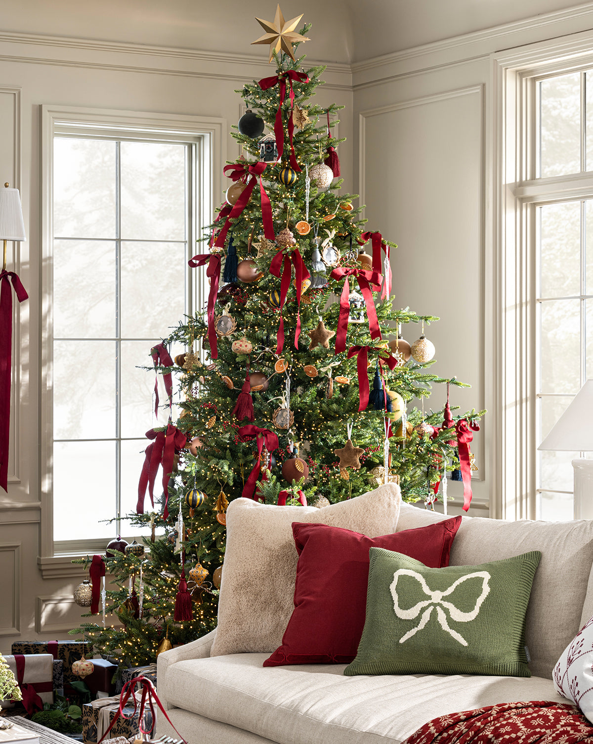 A Christmas tree with red ribbons, ornaments, and a gold star stands by large windows. Wrapped gifts sit below, while a beige sofa topped with red and green pillows plus the Bella Pillow Cover adds cozy holiday charm, by McGee & Co.