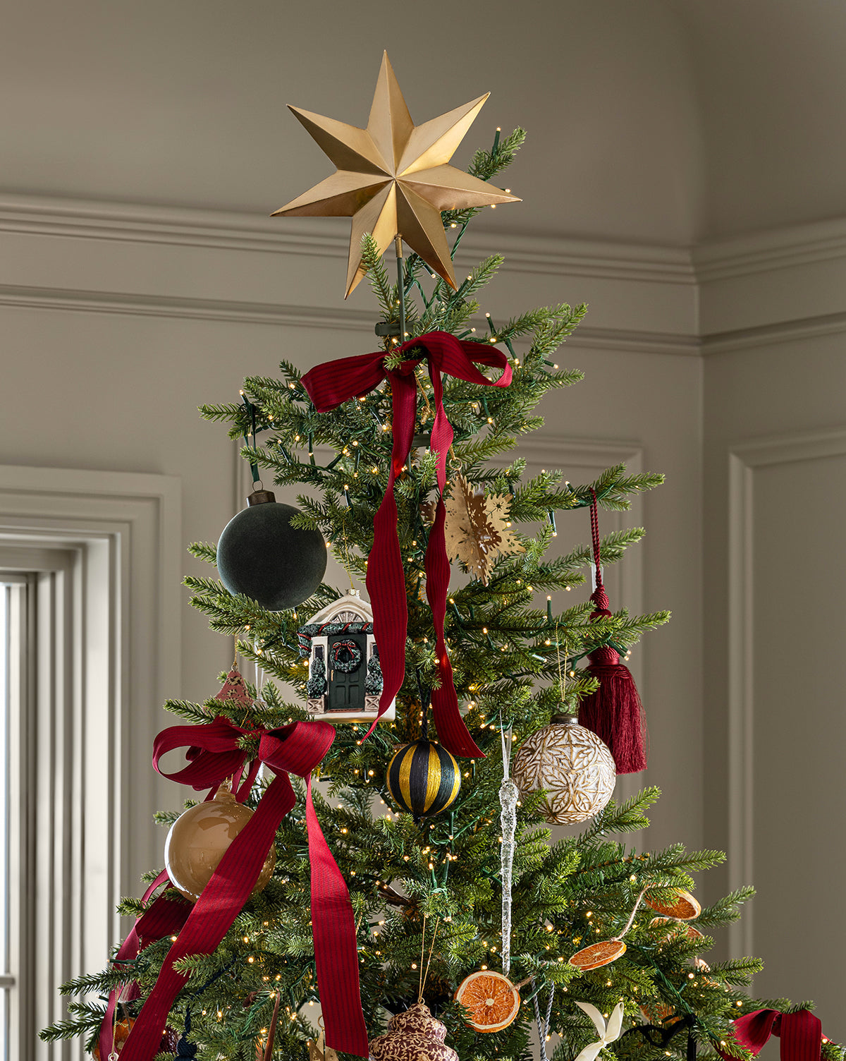 The Classic Christmas Tree is topped with a gold star and decorated with red ribbons, ornaments, dried orange slices, and festive touches. Its setting features light-colored walls and trim for a cozy holiday feel, by McGee & Co.
