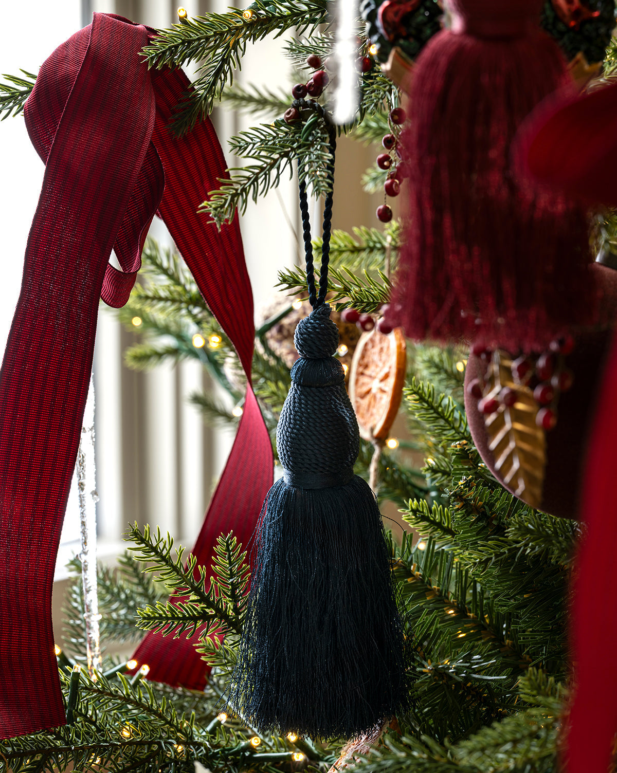 A close-up of a Christmas tree featuring the Lucia Bullion Tassel ornament, accented with a large red ribbon, dried orange slices, berries, and warm white string lights for a touch of vintage-inspired charm, by McGee & Co.