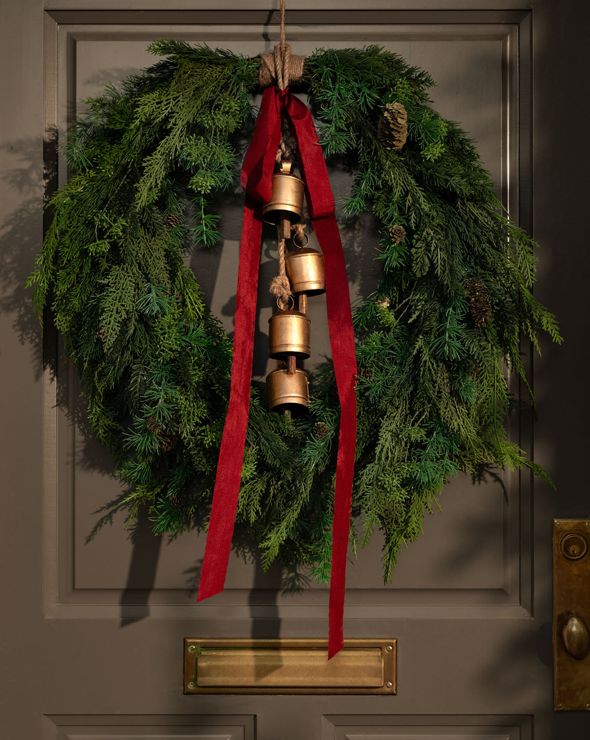 A green holiday wreath with pinecones and a red ribbon hangs on a gray door. Hanging Iron Bells with a gold finish add to the festive décor, while a brass mail slot is visible below, by McGee & Co.