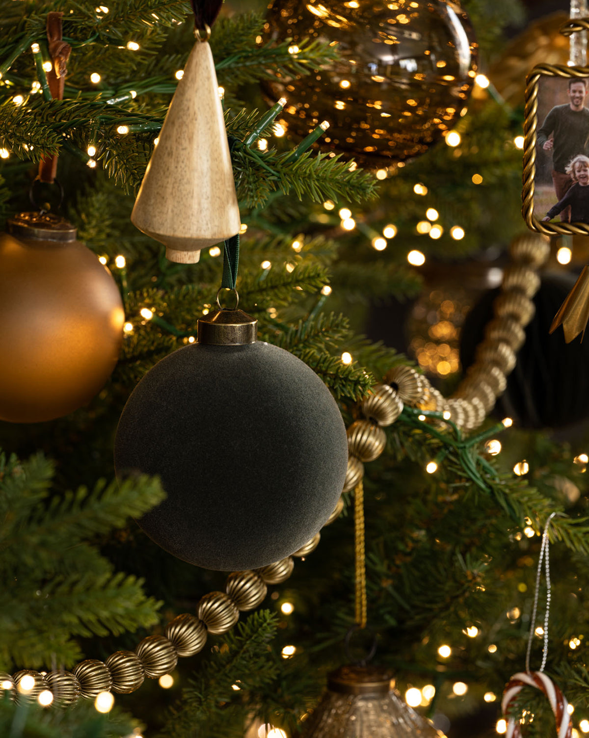 Close-up of a Christmas tree decorated with gold and black ornaments, a velvet black bauble, gold bead garland, warm white string lights, and Flocked Baubles (Set of 8). A small framed photo ornament is partially visible on the right, by McGee & Co.