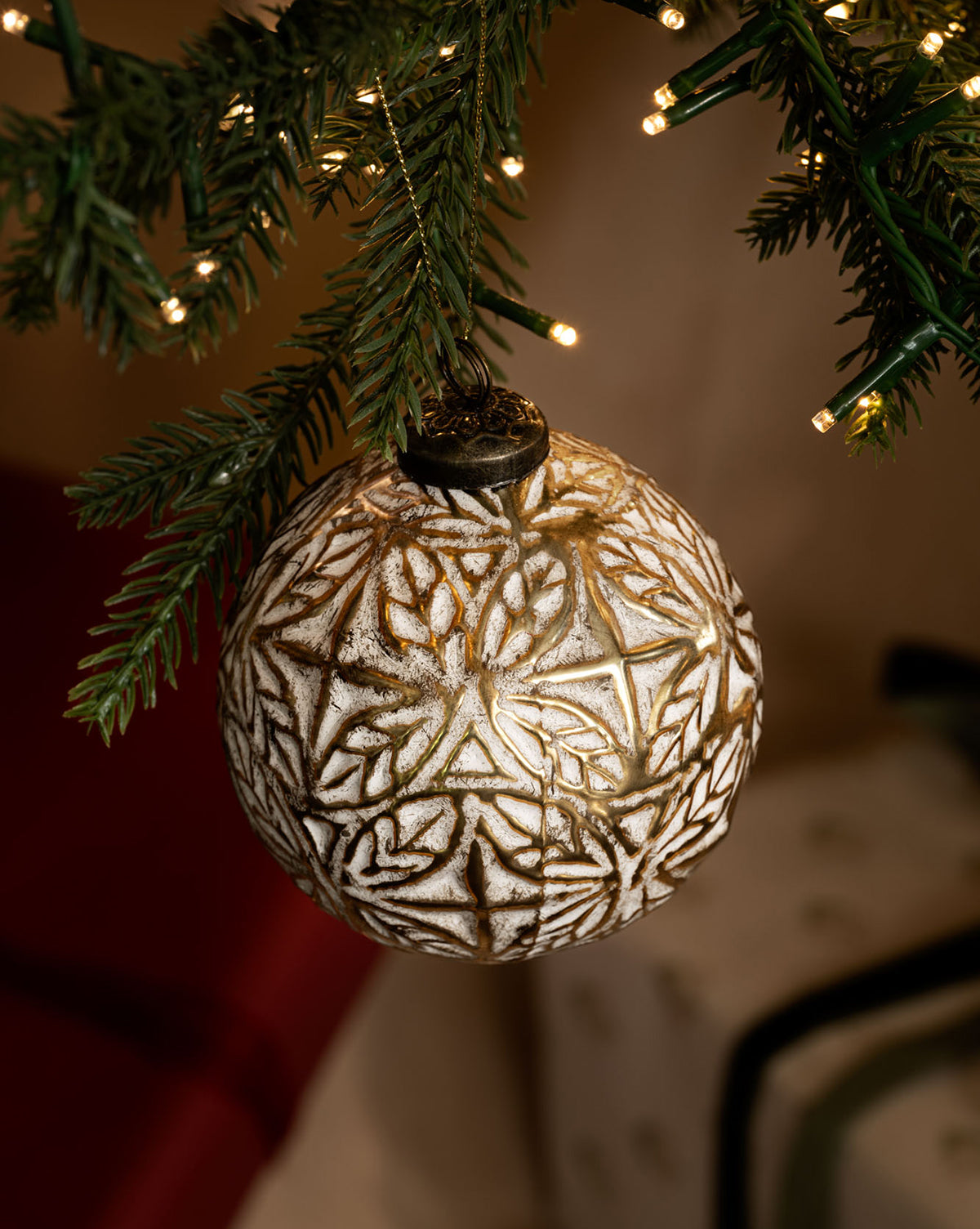 The Alessandro Glass Ornament, featuring a white and gold motif, hangs from a lit evergreen branch with warm string lights and blurred holiday decor in the background, by McGee & Co.