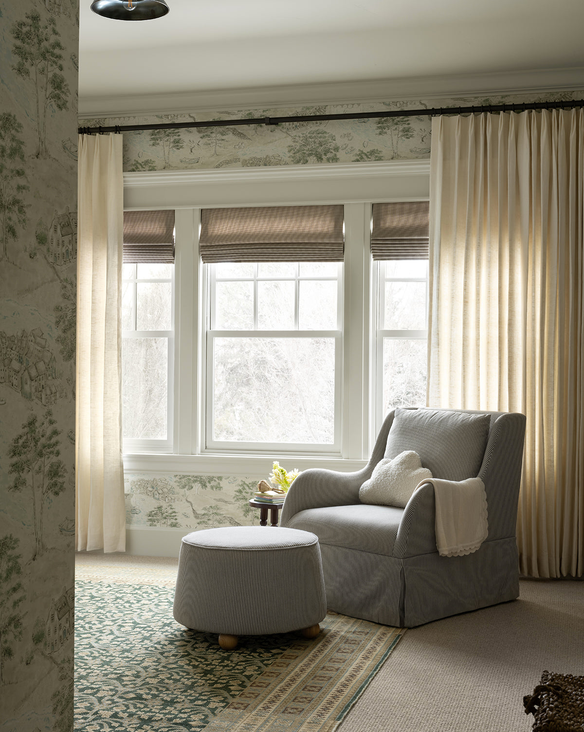 A cozy reading nook with a plush seat, Barnaby Ottoman in gray, a cream throw blanket, green patterned rug, ash wood accents, sheer curtains, and large windows that fill the space with soft natural light, by McGee & Co.