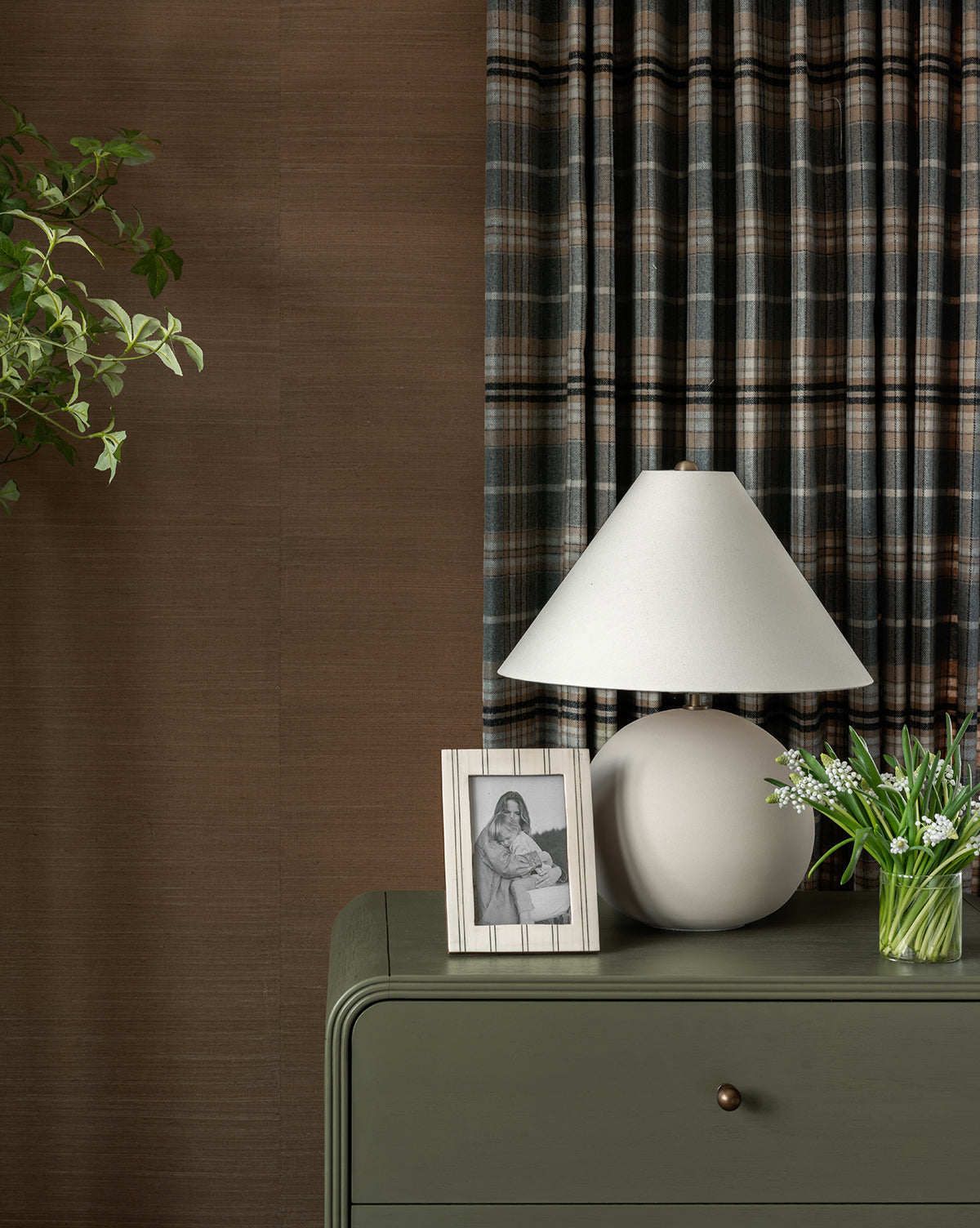 A green dresser topped with the Tess Ceramic Table Lamp, a framed black-and-white photo, and a small vase of flowers stands before plaid curtains and a brown wall. Green plant leaves hang in the upper left corner, by McGee & Co.