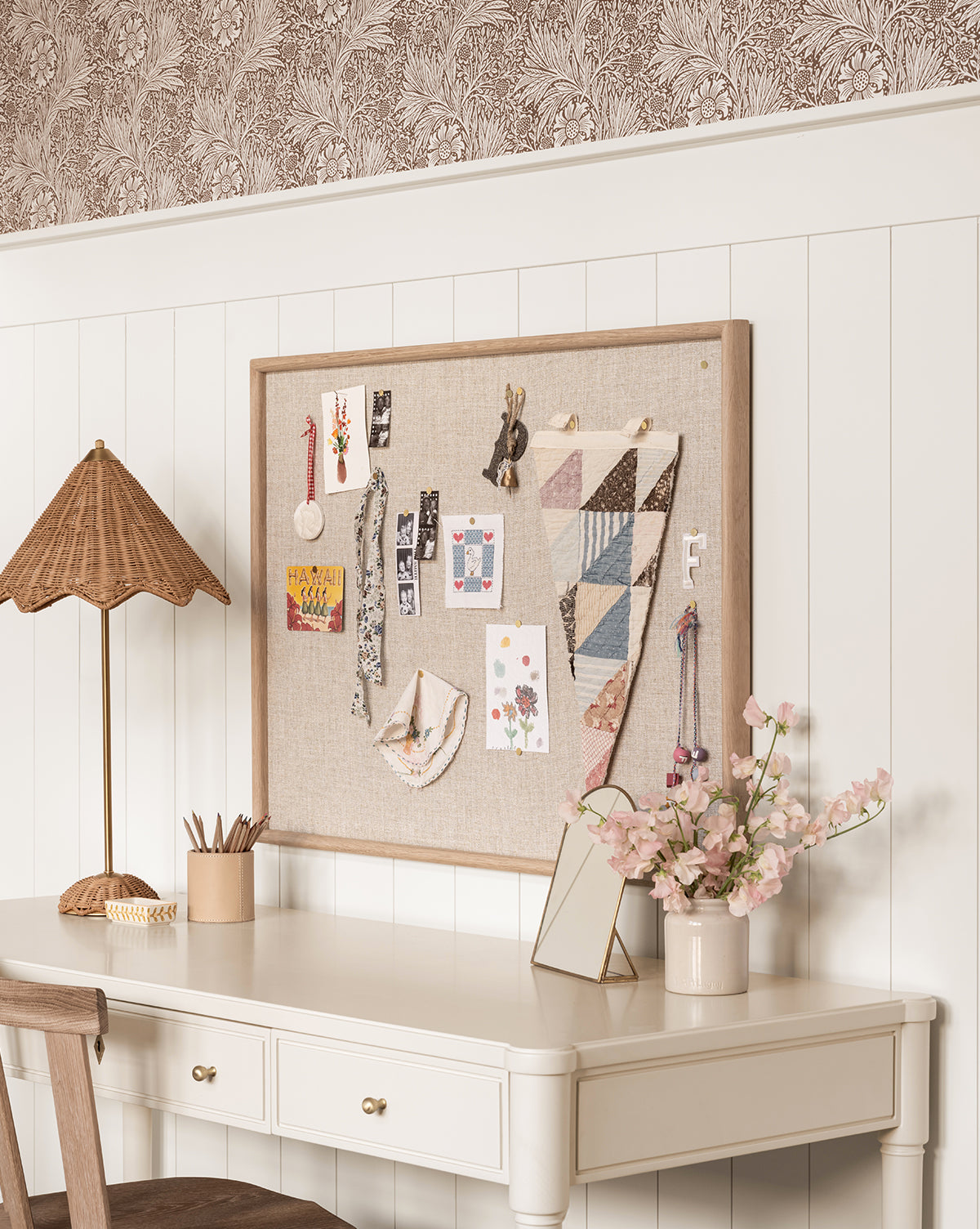 A cream-colored desk with flowers, a lamp, and pencils sits against a white paneled wall. Above, the Oak & Linen Memo Board displays cards, pennants, and crafts. Patterned wallpaper above adds chic organization to the office space.