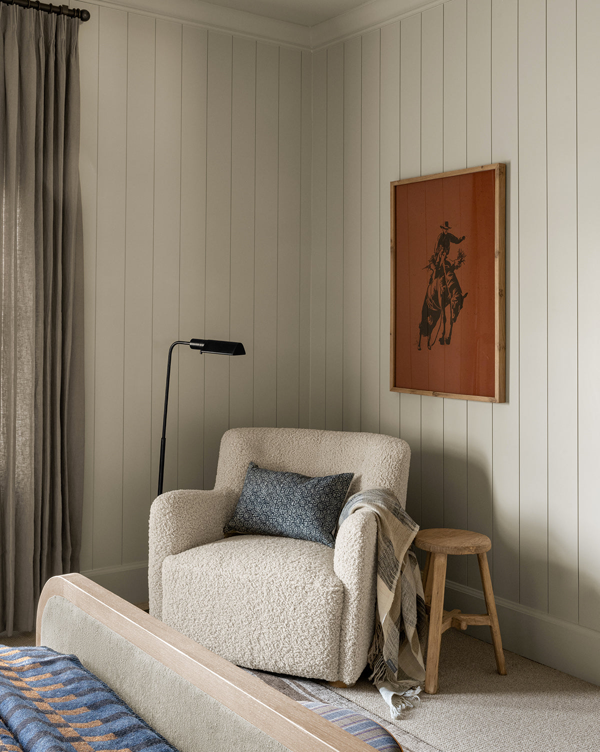 A cozy reading nook features the Magda Lounge Chair (Ready to Ship) with a blue-gray pillow and plaid throw blanket. A wooden stool and black floor lamp are nearby; Western art on white shiplap walls, with gray curtains framing the window, by McGee & Co.