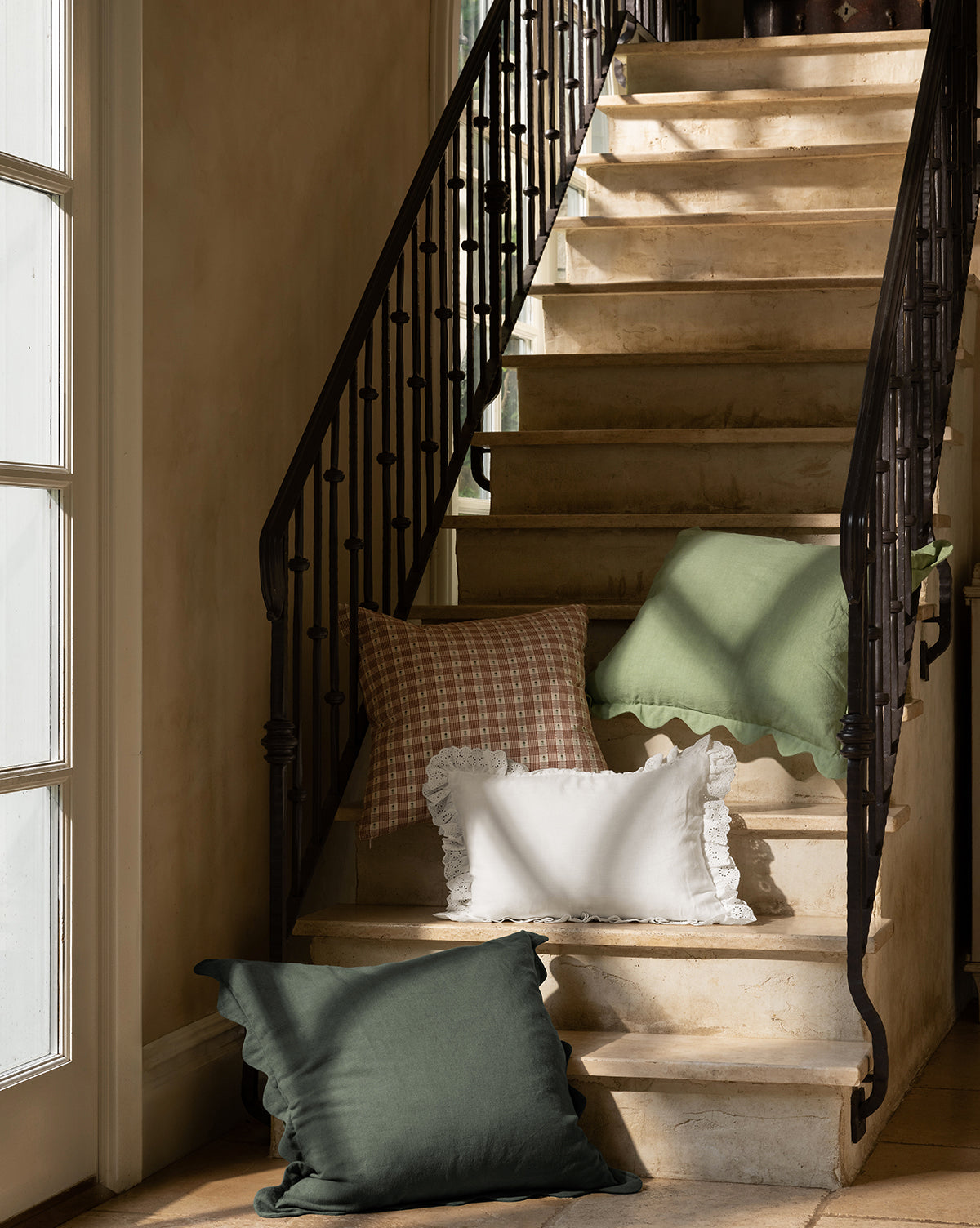 A sunlit stone staircase with a black iron railing is decorated with McGee & Co. x Loeffler Randall Colette Scallop Border Pillow Cover, plus green, ruffled white, and pink plaid pillows. Light from the left window highlights their cozy mix of textures, by McGee & Co.